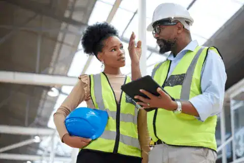 Two construction workers wearing safety vests and helmets have a discussion indoors.